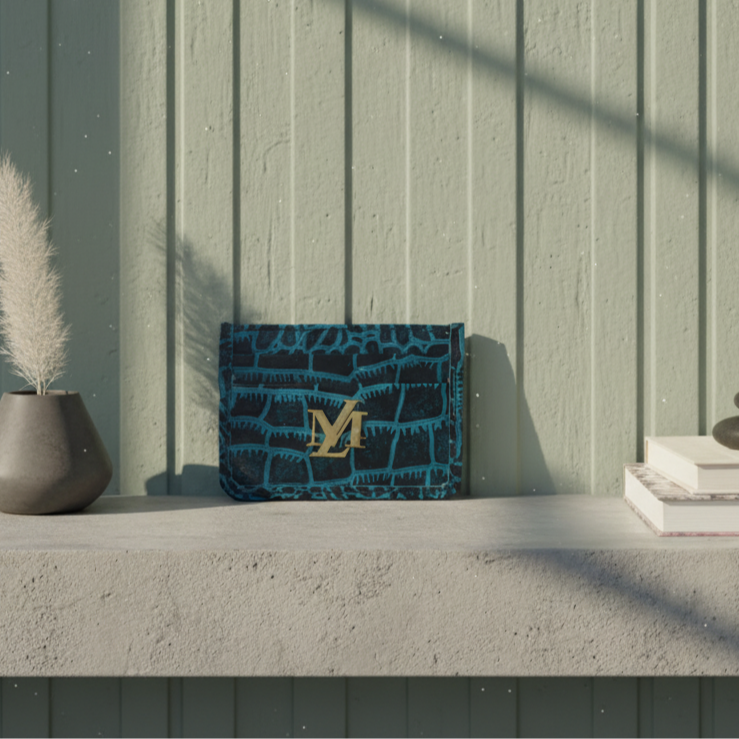 Blue crocodile-patterned handbag with gold logo on a concrete surface against a light green wooden wall.